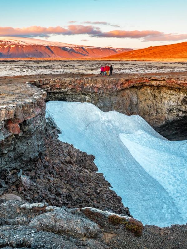 A deep cave opening in dark volcanic rock, partially filled with snow, with people at the rim and mountains under a golden sky.