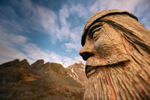 Rostro tallado en madera de un hombre barbudo mirando sobre montañas cubiertas de nieve bajo un cielo azul nublado.
