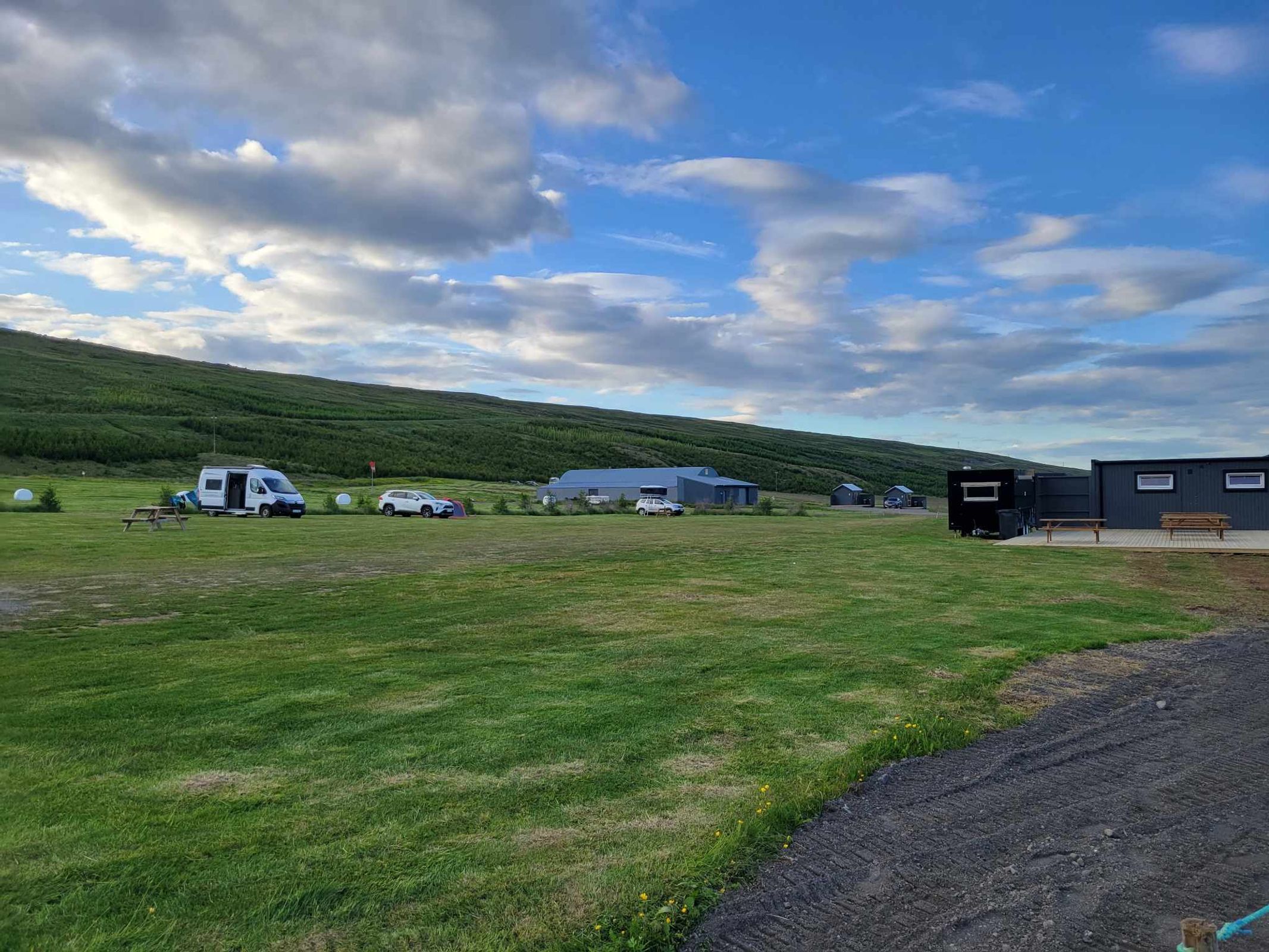 A grassy campsite with a camper van, cars, and modular cabins, set against a green hill under a partly cloudy sky.