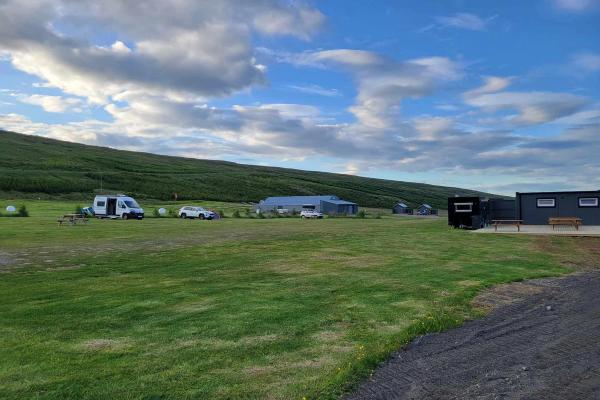 A grassy campsite with a camper van, cars, and modular cabins, set against a green hill under a partly cloudy sky.
