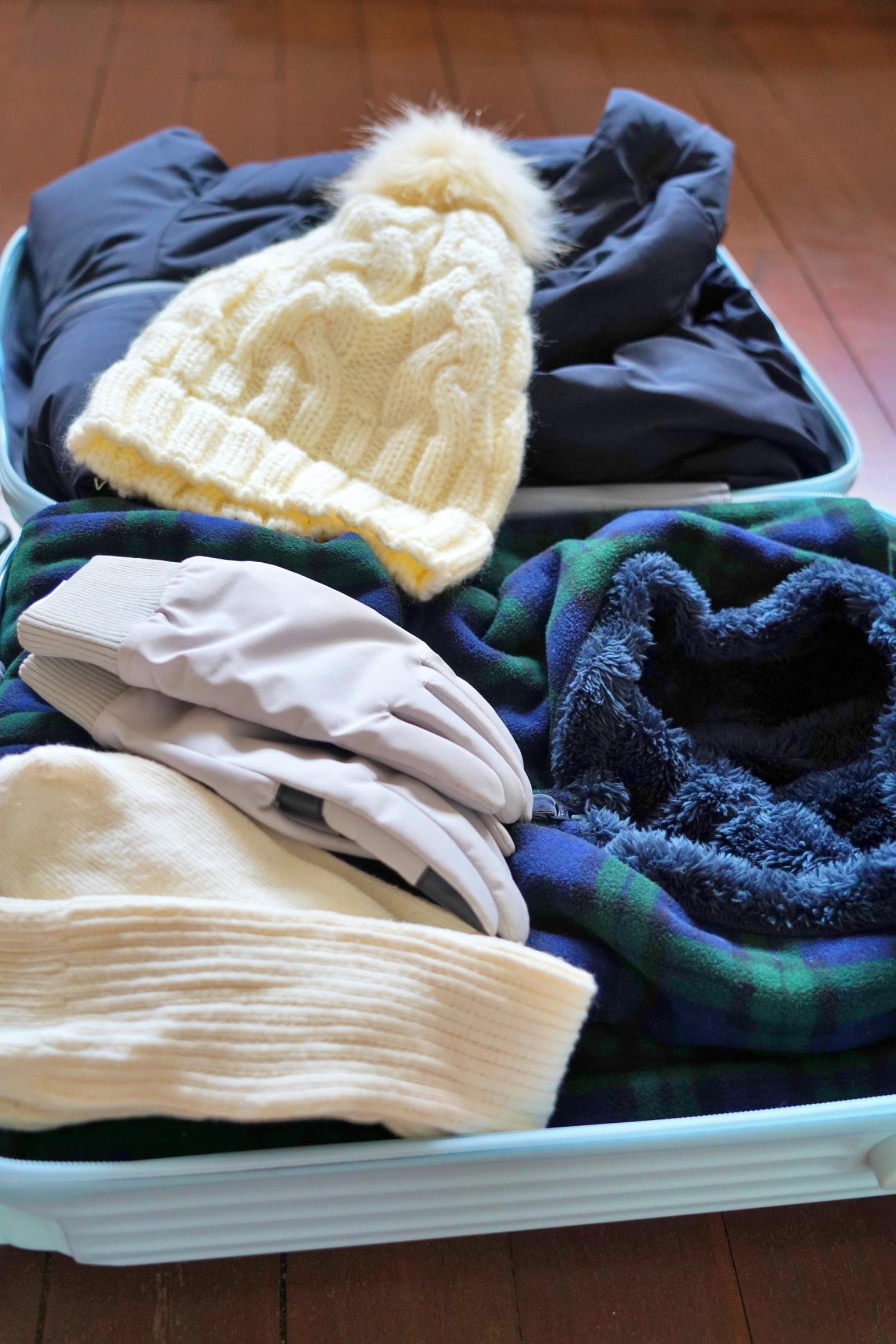 Winter clothing including a cream knitted hat, grey gloves, a dark blue jacket, and a plaid fleece blanket packed in a light blue suitcase.