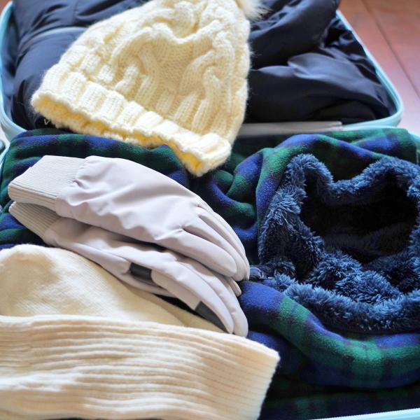 Winter clothing including a cream knitted hat, grey gloves, a dark blue jacket, and a plaid fleece blanket packed in a light blue suitcase.
