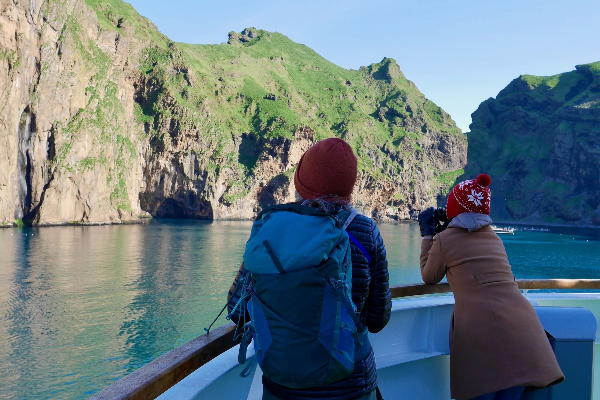 Two people on a boat look at towering green cliffs with a waterfall and sea caves, one holding binoculars.