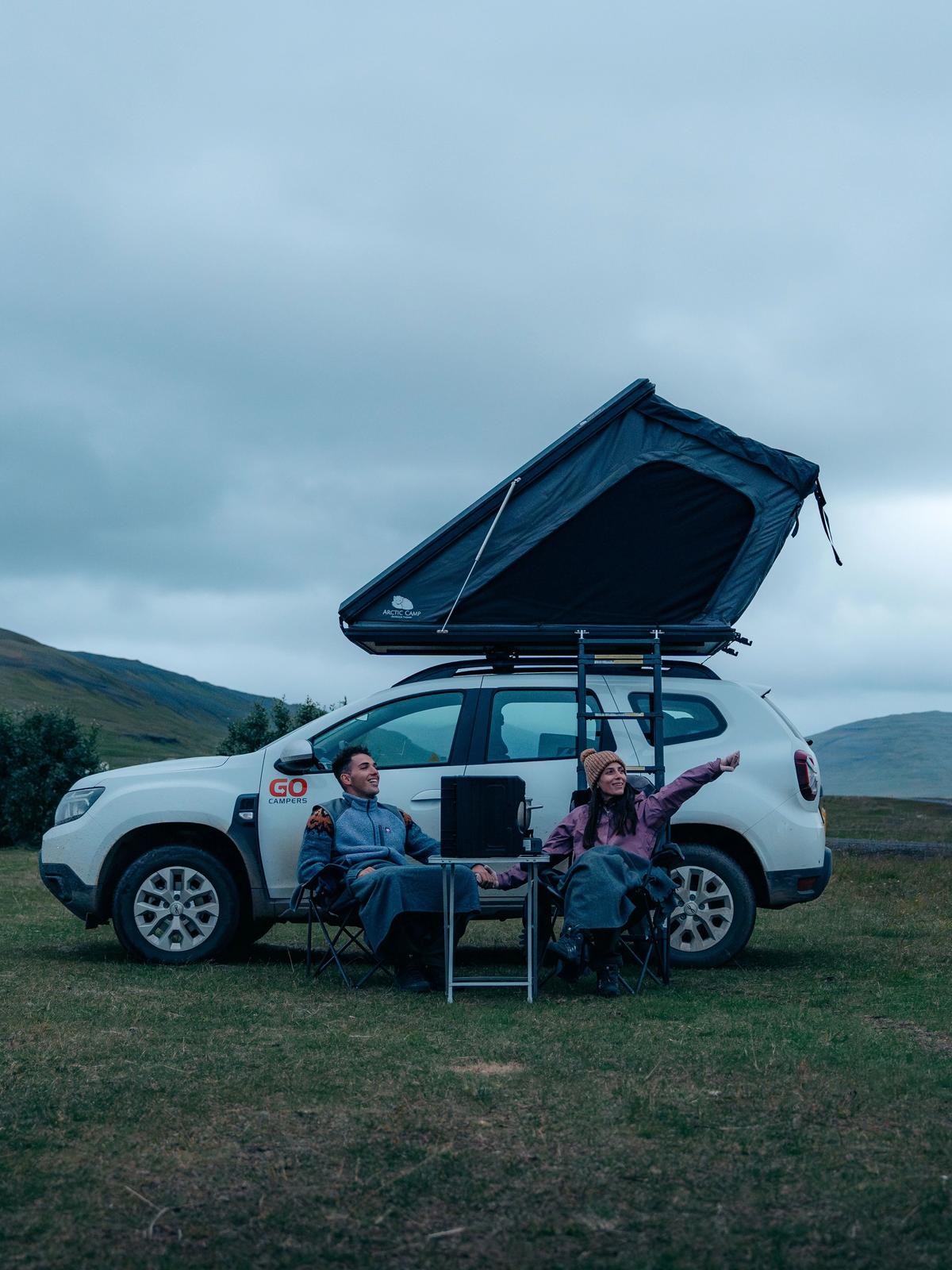 Two people sit with a laptop next to a white SUV with an open rooftop tent in a grassy field; a woman points towards distant hills under a cloudy sky.