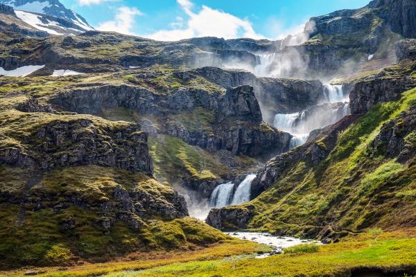 Terraced waterfalls cascade down a green, rocky mountain into a stream, with mist rising.