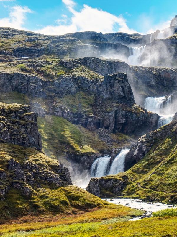 Cascading waterfalls flow down a green, rocky mountain, enveloped in mist, under a blue sky.