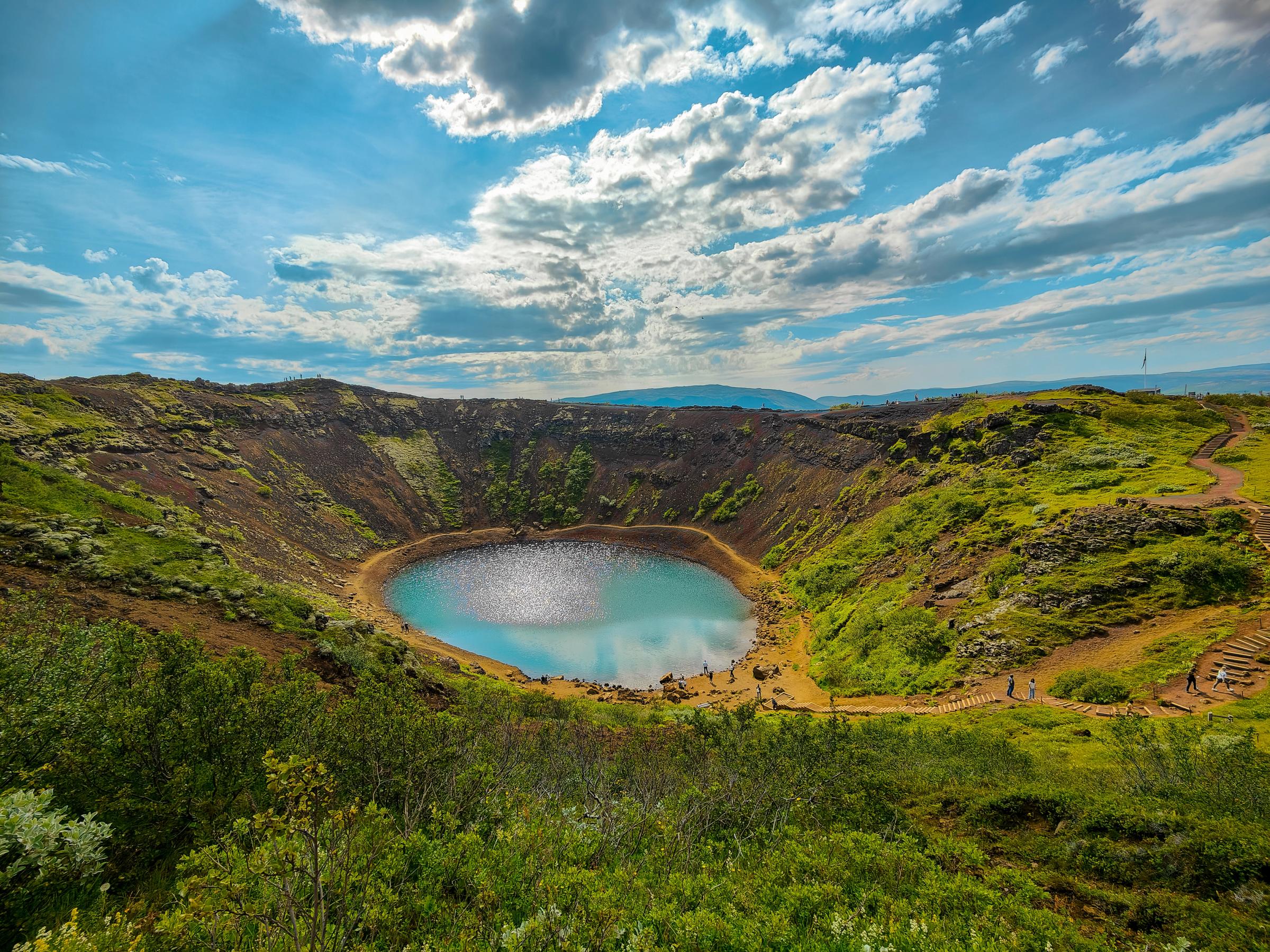 Volcanic crater with a bright blue lake surrounded by green slopes under a cloudy sky.