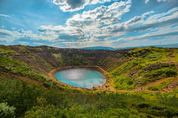 A vibrant blue crater lake surrounded by reddish-brown earth and green vegetation under a partly cloudy sky, with people walking along a path on the rim.