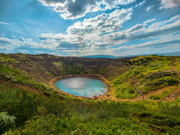 Kerid volcanic crater with a vibrant blue-green lake, surrounded by green and brown slopes, under a partly cloudy blue sky.