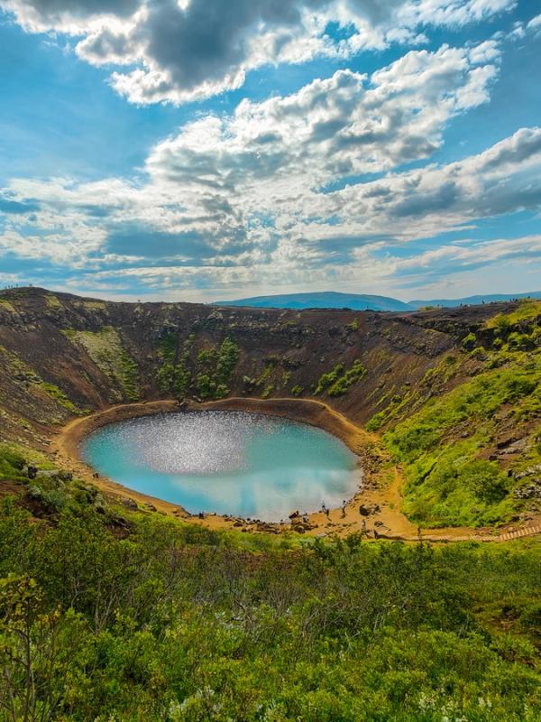 A volcanic crater with a vibrant blue lake, green slopes, and a winding path.