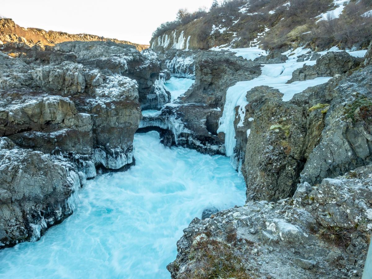 A bright turquoise river flows through a rocky, snow-covered canyon with ice formations.