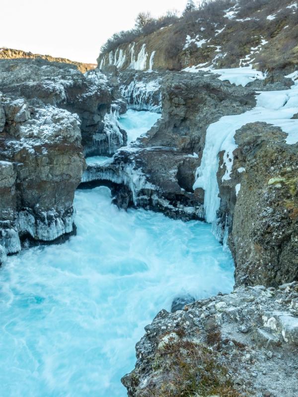 A bright turquoise river flows through a rocky, snow-covered canyon with ice formations.
