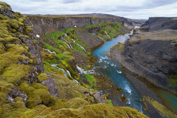 A vibrant blue river flows through a canyon, with lush green moss and waterfalls on one side and dark volcanic rock on the other.
