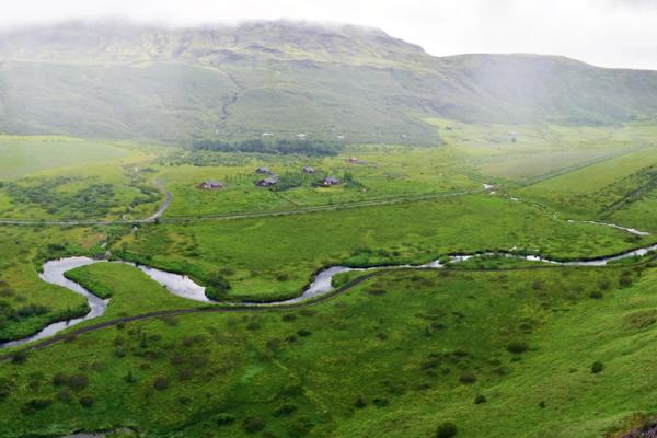 A panoramic view of a lush green valley with a winding river, distant buildings, and cloud-covered mountains.