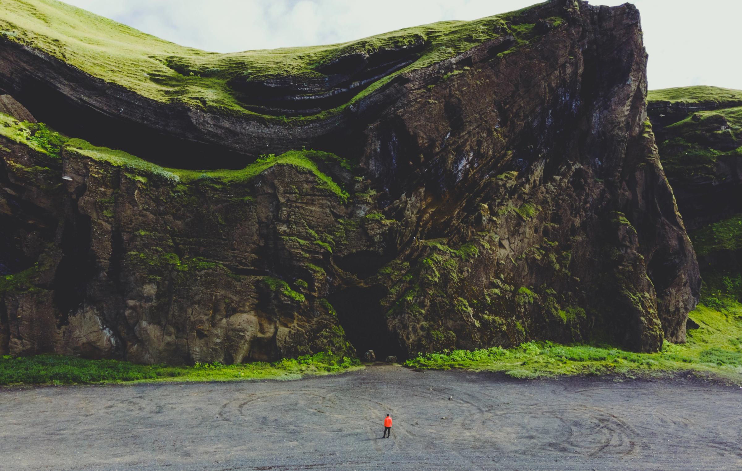 A tiny person in an orange jacket stands on a dark beach before a massive, layered cliff with green, mossy slopes.