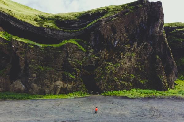 A person in an orange jacket stands on a dark beach before a towering, moss-covered cliff.