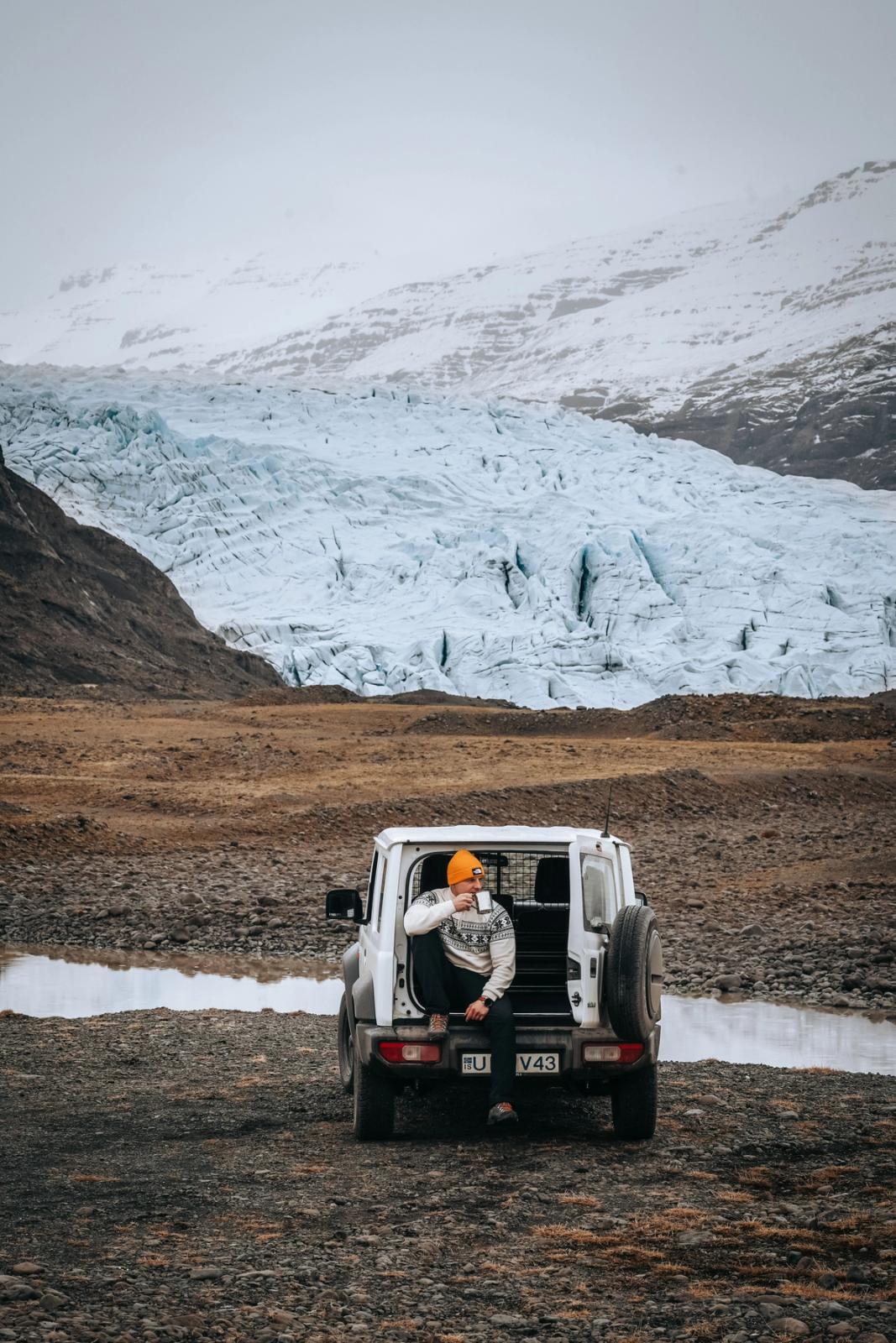 A person in an orange hat sits in the open trunk of a white SUV, drinking from a mug, overlooking a large glacier and snowy mountains.