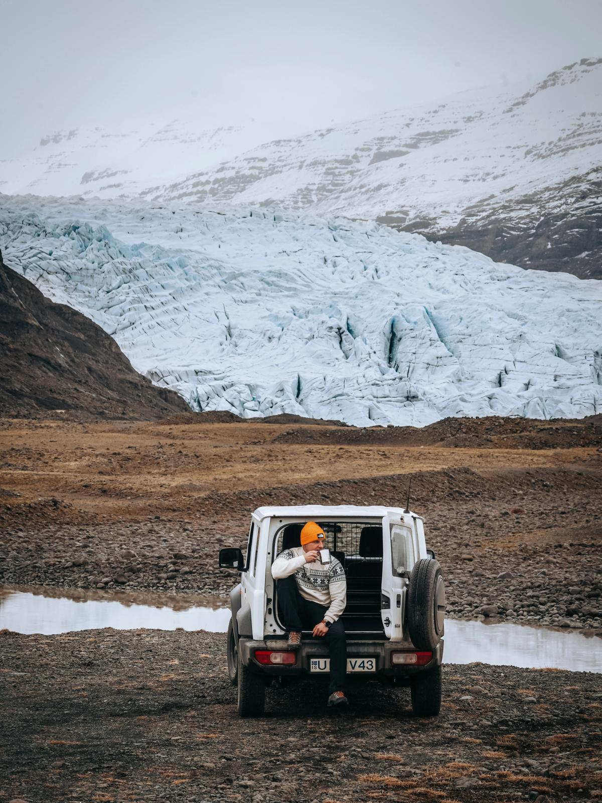 A person in an orange hat sits in the open trunk of a white SUV, drinking from a mug, overlooking a large glacier and snowy mountains.