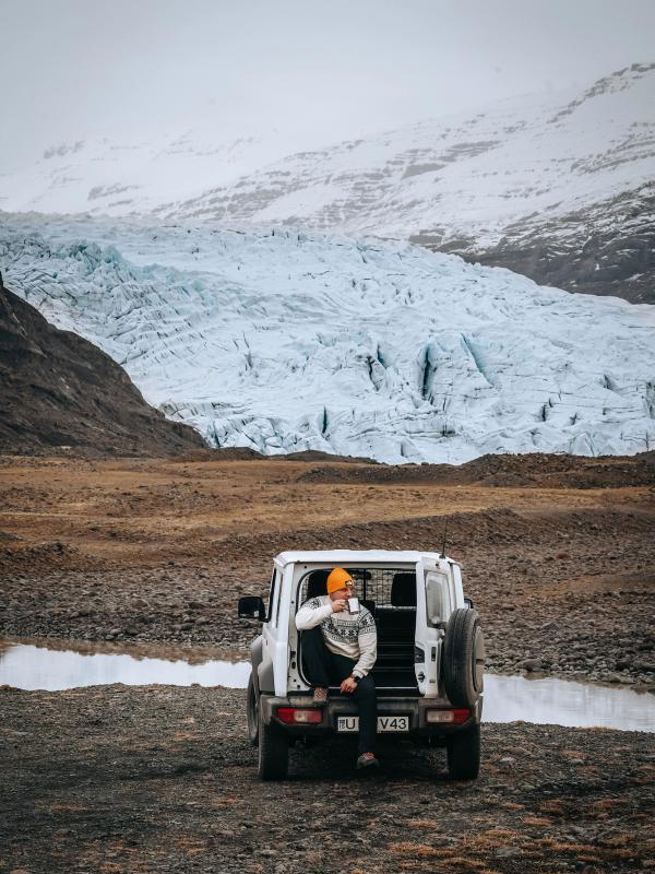 A person in an orange hat sits in the open trunk of a white SUV, drinking from a mug, overlooking a large glacier and snowy mountains.