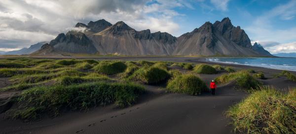 Una persona con una chaqueta roja camina por una playa de arena negra con mechones de hierba verde, hacia montañas escarpadas oscuras y el océano.