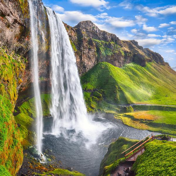 Tall waterfall flowing down a green mossy cliff, surrounded by vibrant green hills and a small wooden bridge, under a bright blue sky.