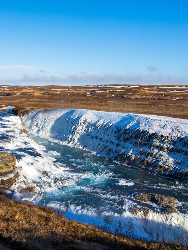 A powerful waterfall cascades through a snowy, barren landscape under a clear blue sky.