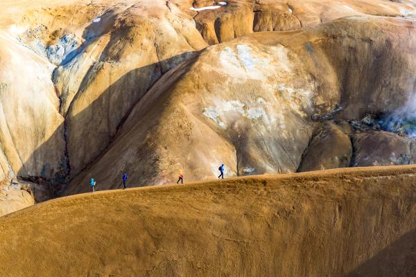 A line of hikers walks along a ridge in a brown and white geothermal landscape with steam.