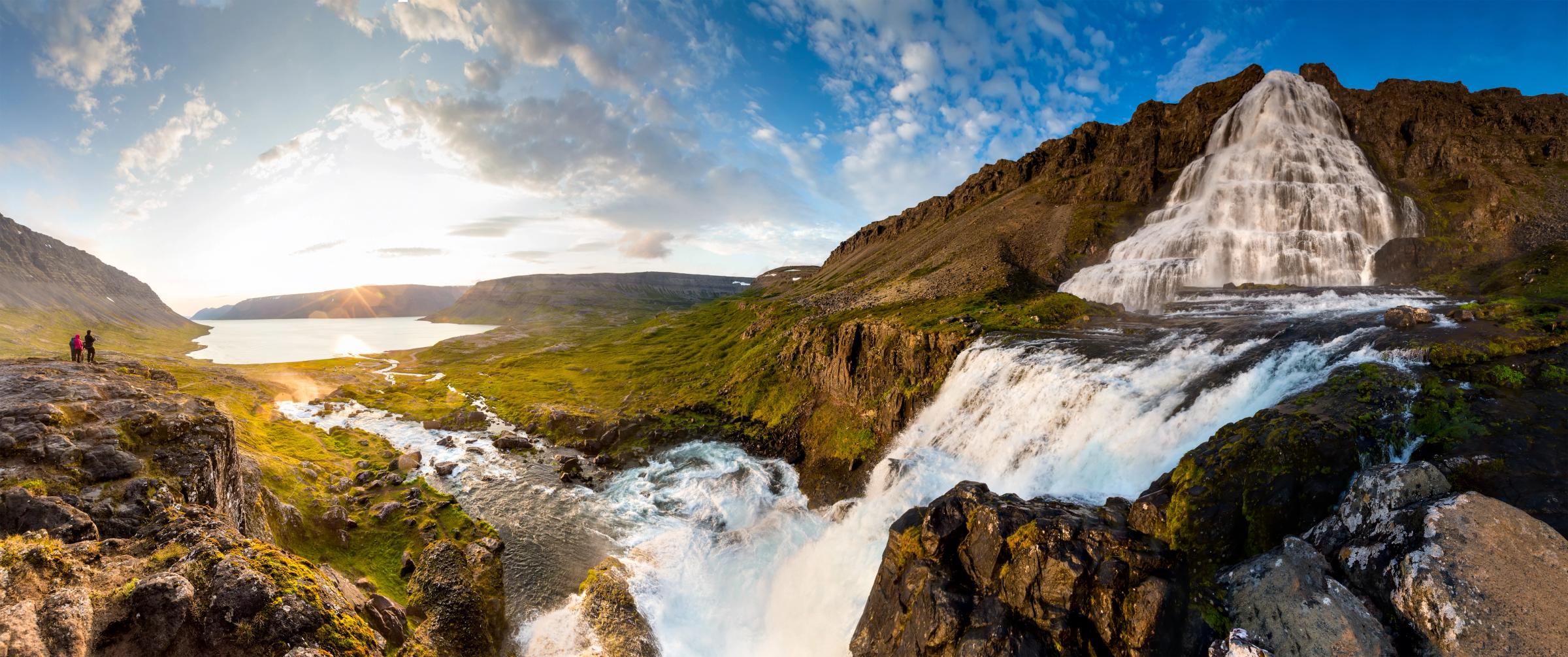 Vista panorámica de la cascada escalonada Dynjandi, que cae en una verde valle con un fiordo y un atardecer de fondo.