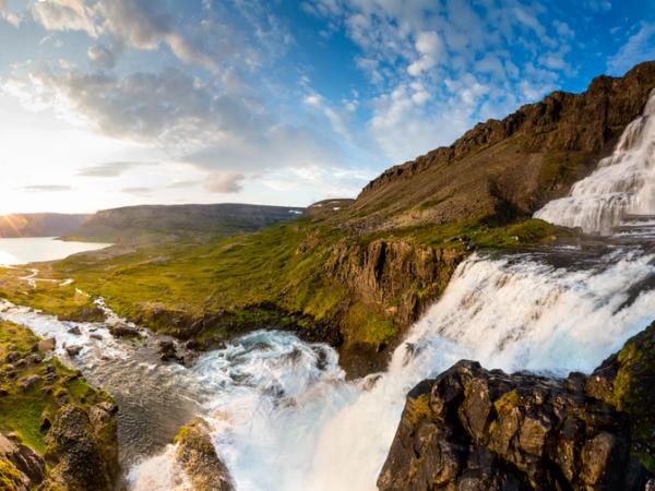 Vista panorámica de la cascada escalonada Dynjandi, que cae en una verde valle con un fiordo y un atardecer de fondo.