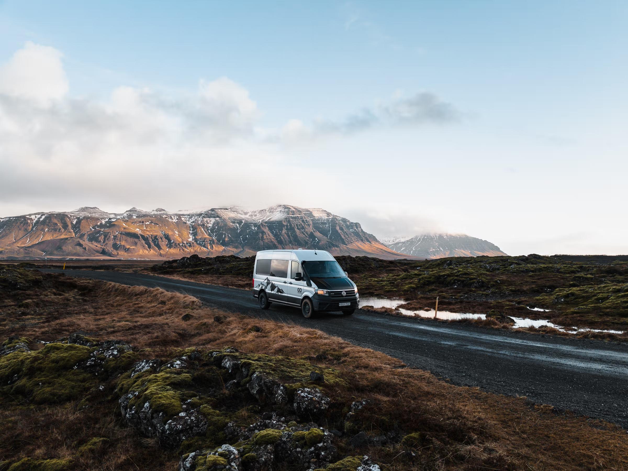 A white camper van drives on a dirt road through a rugged, mossy landscape with snow-capped mountains.