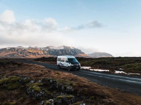 White camper van on a gravel road through a rugged, mossy landscape with snow-capped mountains.
