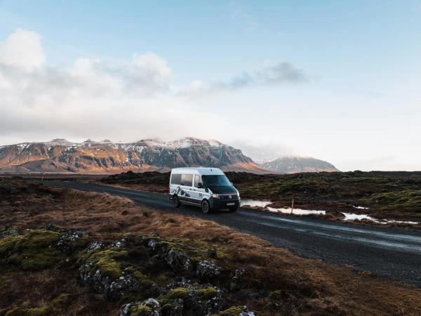 White camper van on a gravel road through a rugged, mossy landscape with snow-capped mountains.