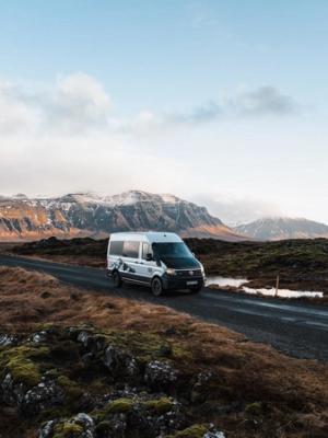 White camper van on a gravel road through a rugged, mossy landscape with snow-capped mountains.