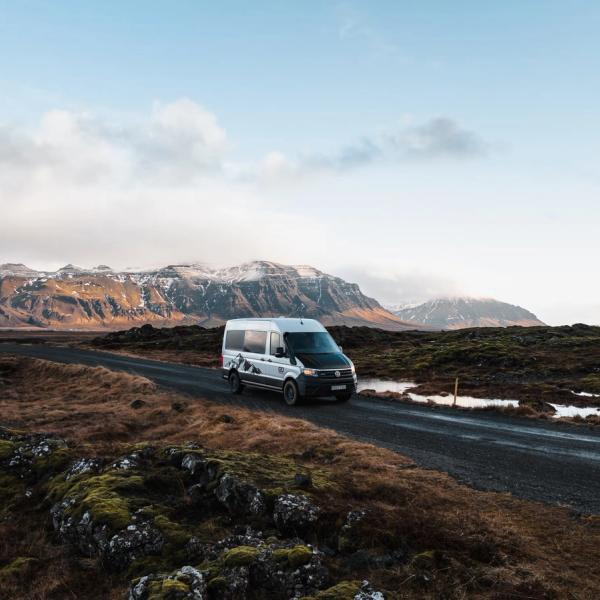 A white camper van drives on a dirt road through a rugged, mossy landscape with snow-capped mountains.