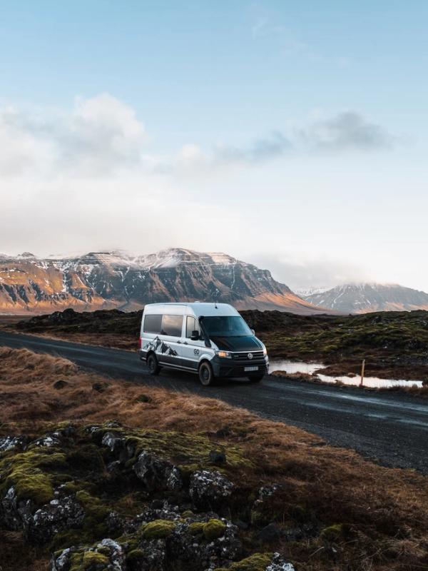 White camper van on a gravel road through a rugged, mossy landscape with snow-capped mountains.