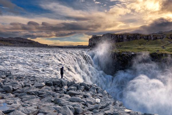 a man is standing on a rocky cliff overlooking Dettifoss waterfall .