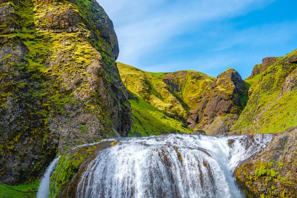 A wide waterfall cascades into a rocky river, surrounded by steep, moss-covered hills under a clear blue sky.