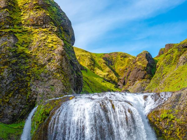 A wide waterfall cascades into a rocky river, surrounded by moss-covered green hills under a bright blue sky.