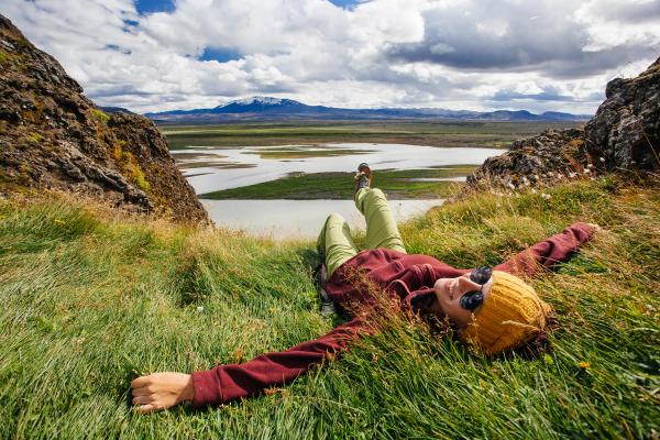 Person in a yellow beanie and sunglasses relaxing on a grassy hill, overlooking a lake and snowy mountains.
