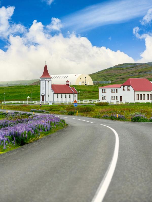 A winding road lined with purple flowers leads to a white church and houses with red roofs in a green, hilly landscape under a blue, cloudy sky.