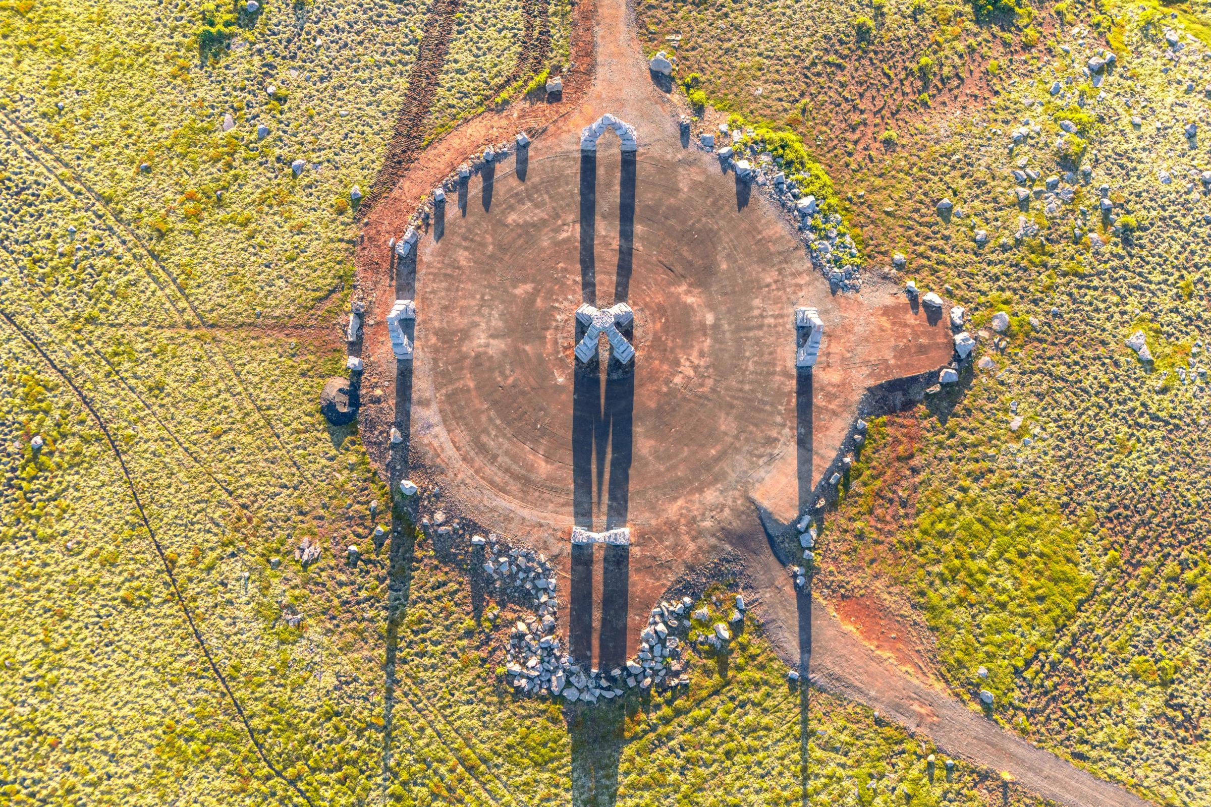 Vista aérea de cuatro estructuras de piedra proyectando largas sombras en forma de cruz sobre un campo circular de tierra rodeado de terreno verde.