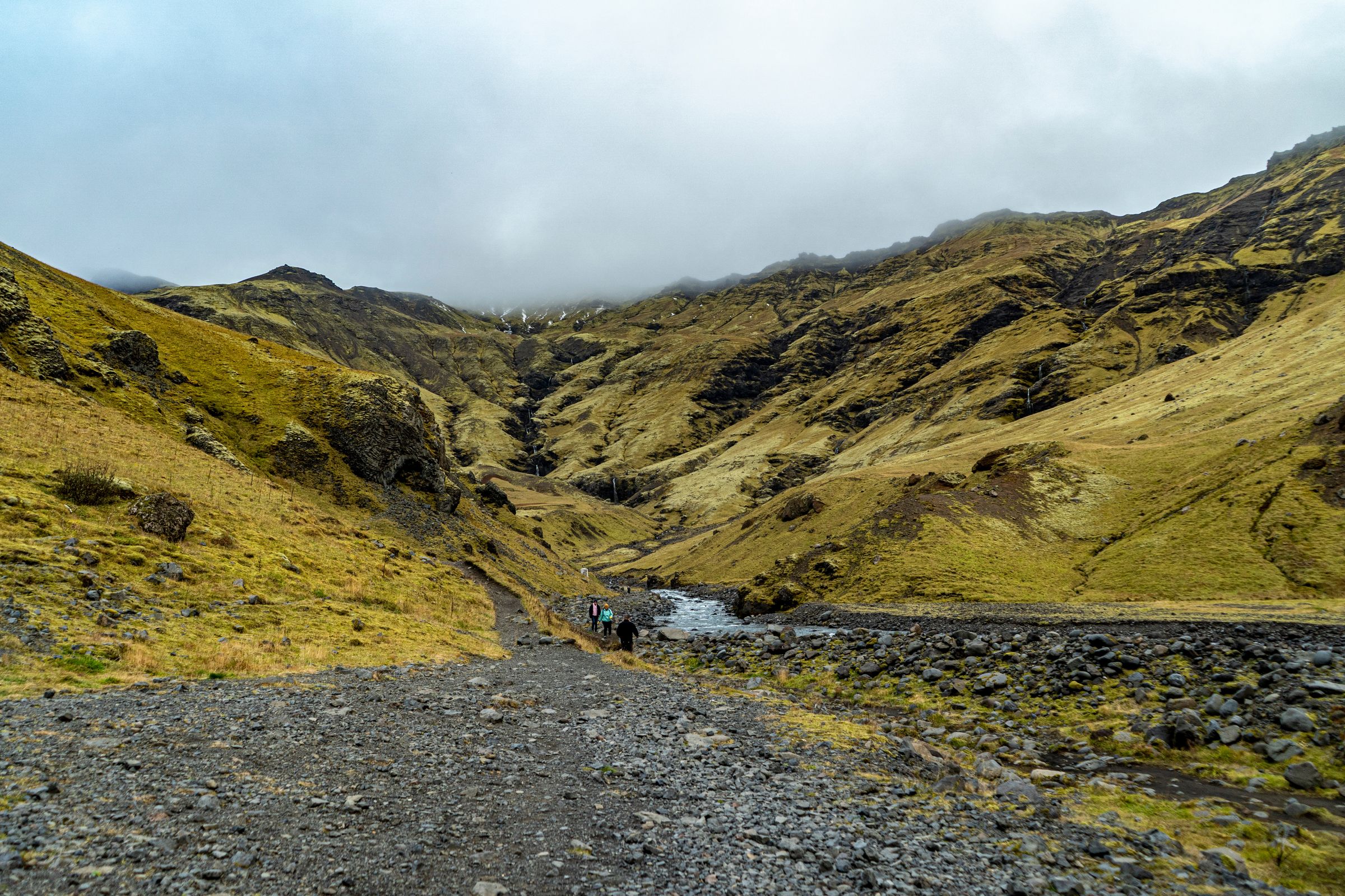 a group of people are walking down a dirt road in the mountains to Seljavallalaug Hot Pool
