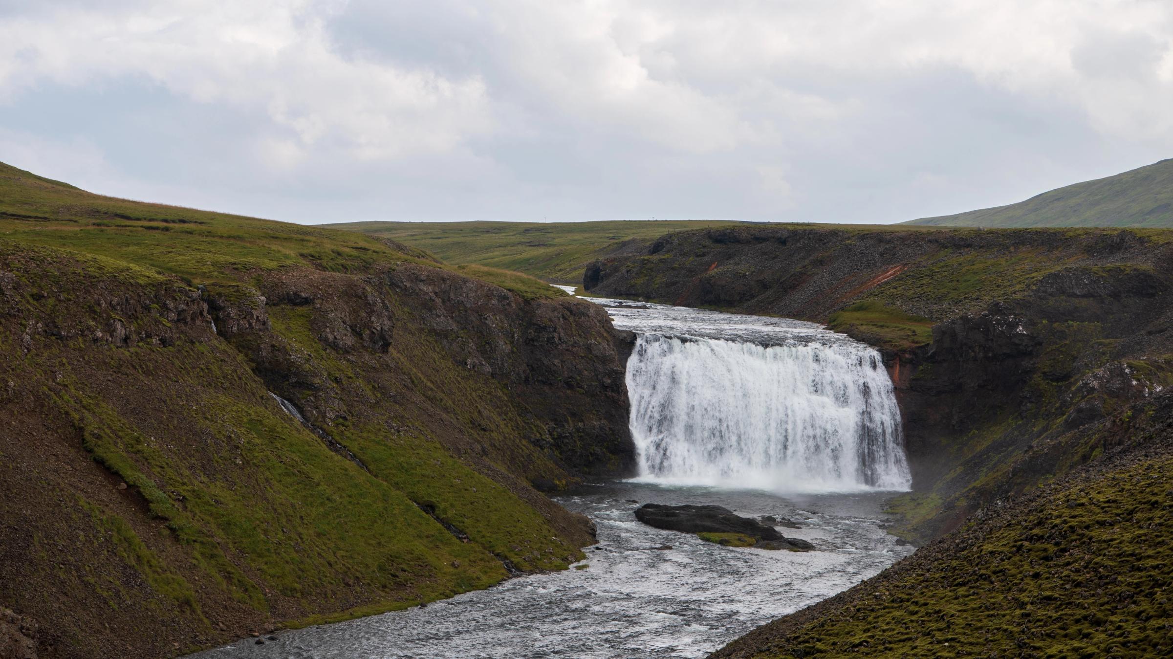 A wide waterfall flows into a river through a green, rocky valley under a cloudy sky.