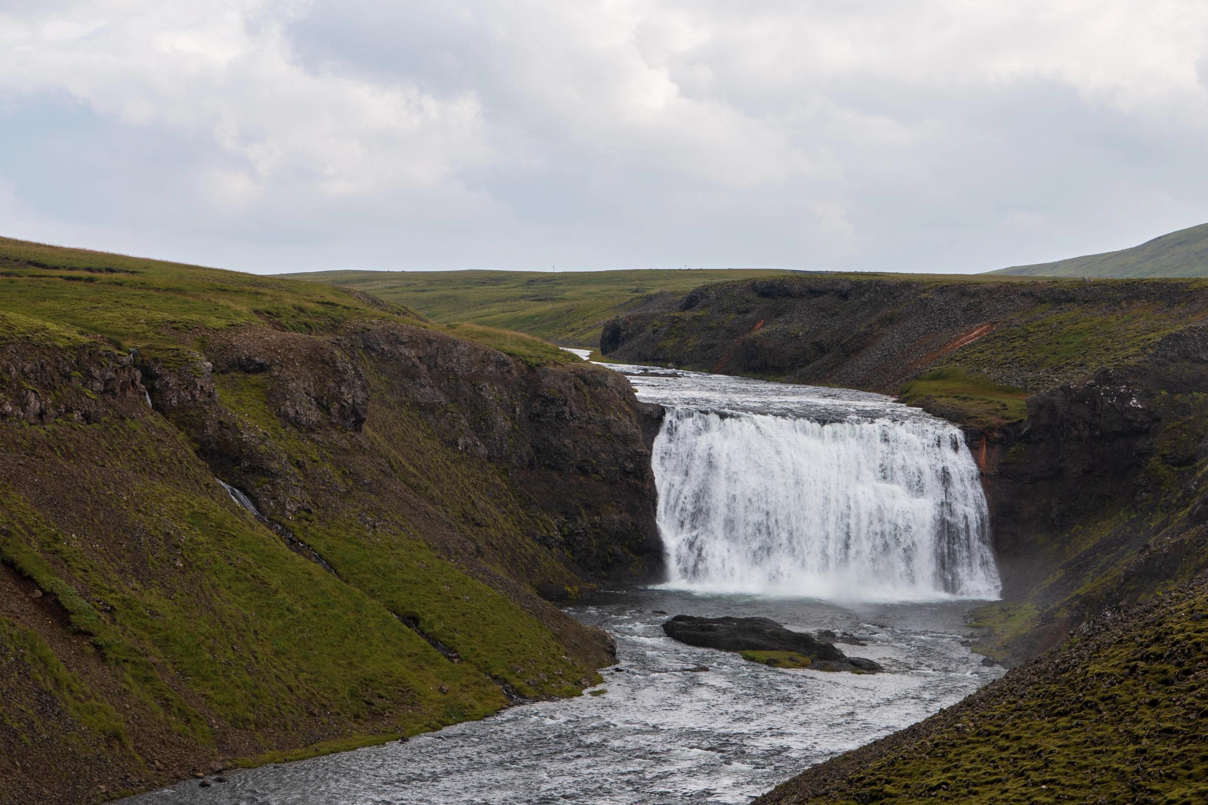 A wide waterfall flows into a river through a green, rocky valley under a cloudy sky.