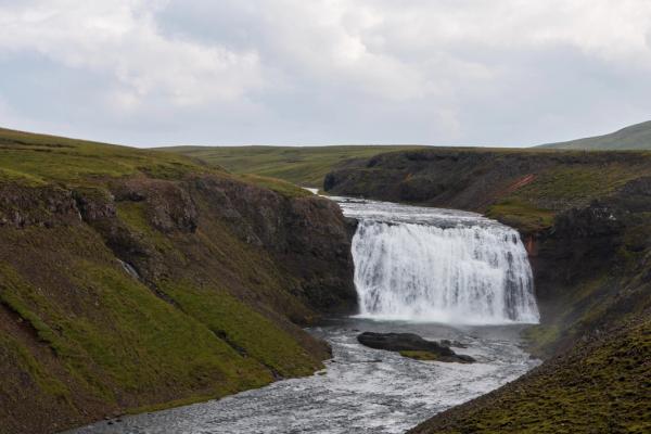 A wide waterfall flows into a river through a green, rocky valley under a cloudy sky.