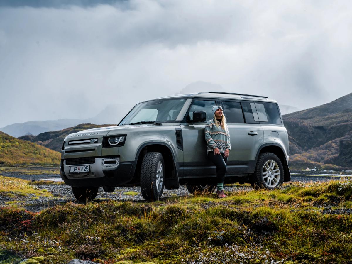 Defensor del paso de montaña Land Rover Defender 4x4 de alquiler en Islandia, aparcado en un terreno accidentado con una persona apoyada en él