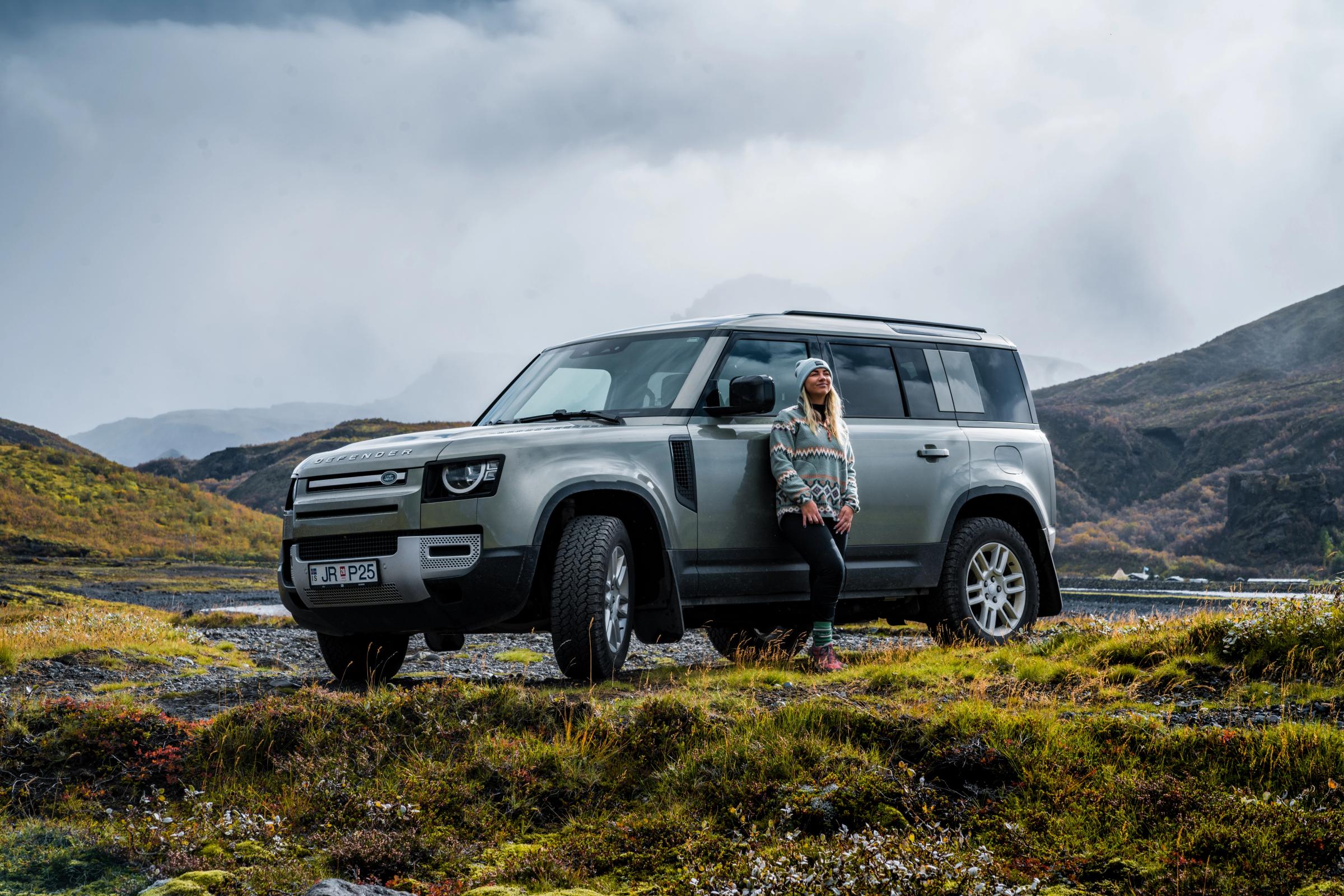 Defensor del paso de montaña Land Rover Defender 4x4 de alquiler en Islandia, aparcado en un terreno accidentado con una persona apoyada en él