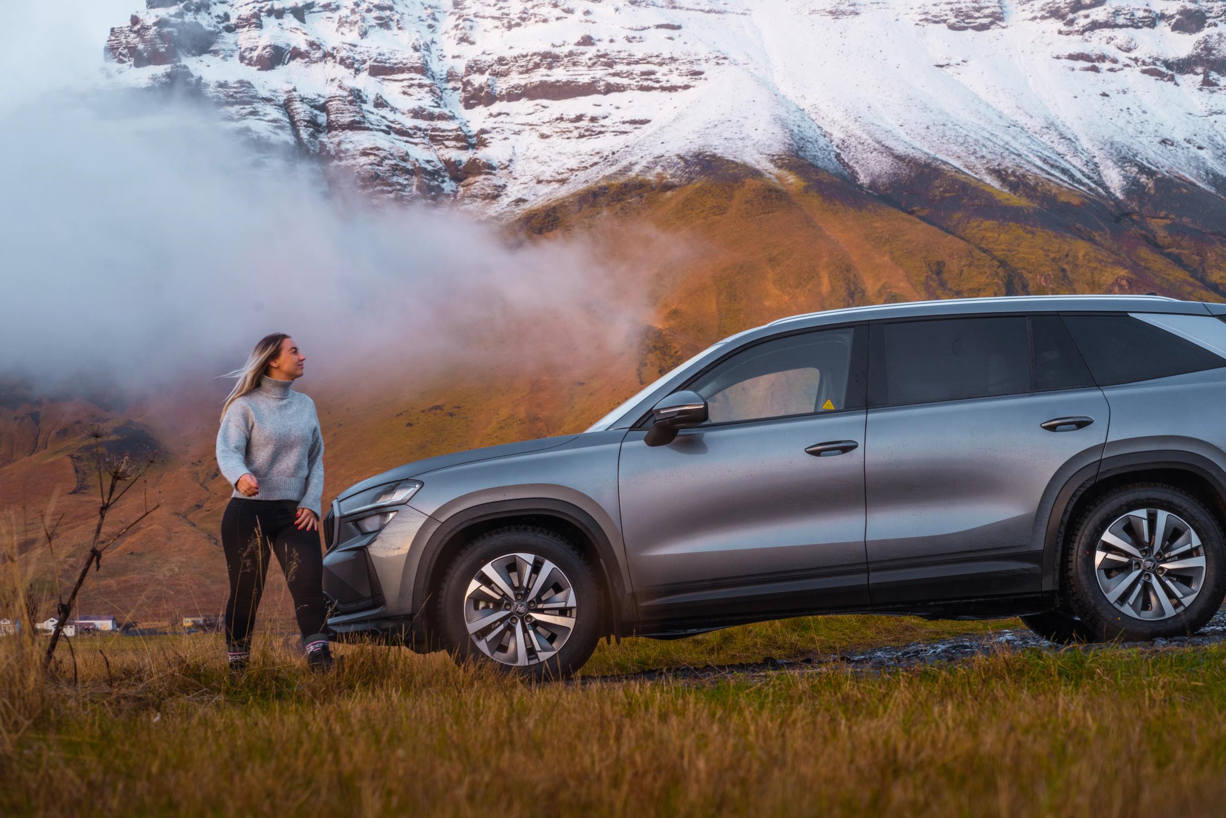 Vista lateral de um Skoda Kodiaq estacionado na relva, com uma pessoa ao lado e montanhas cobertas de neve ao fundo