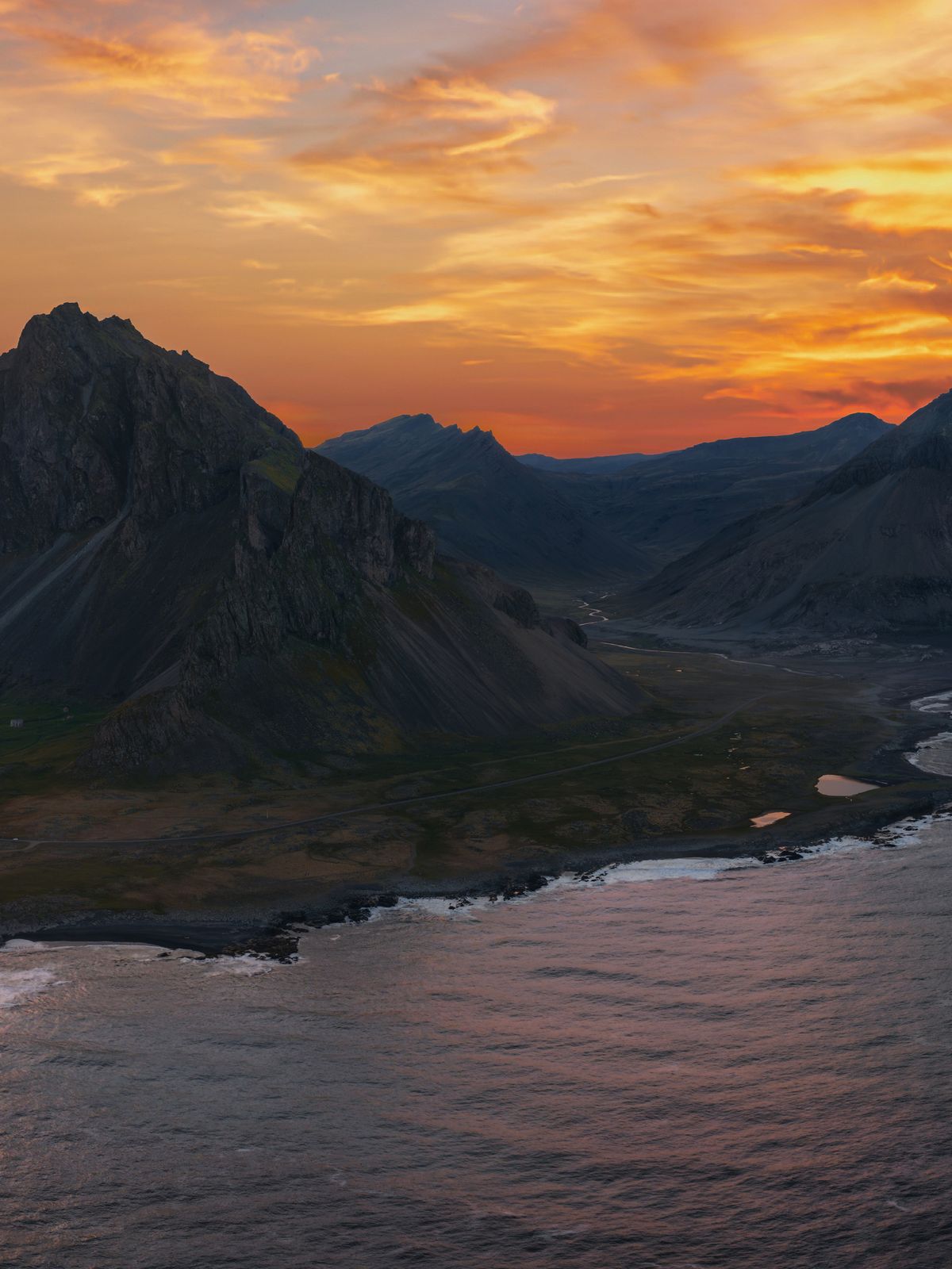 Jagged mountains meet a dramatic coastline and calm bay under a vibrant orange sunset.