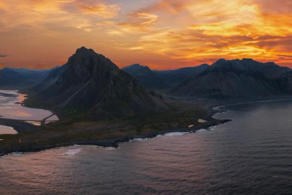 Aerial view of a rugged mountain range on a peninsula with a lagoon and ocean under a vibrant orange and red sunset sky.